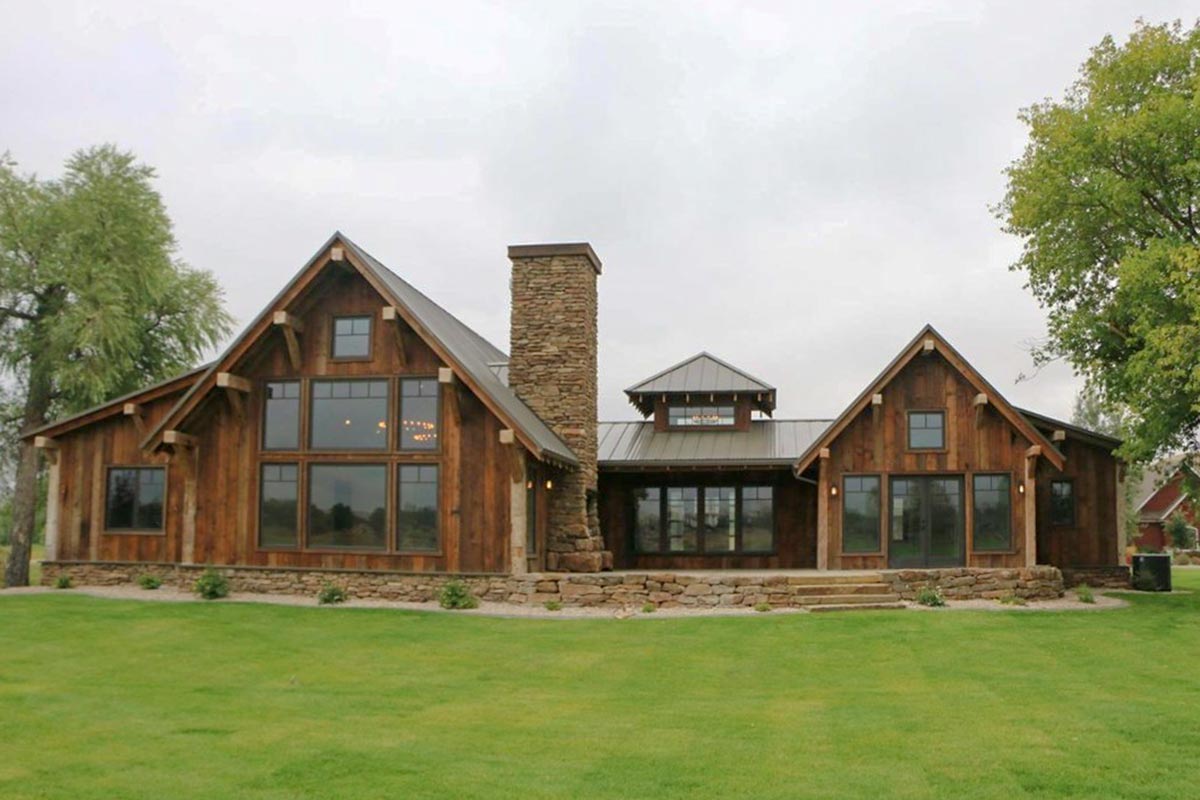 Rear of rustic mountain ranch home with walls of windows, timber gables and stone chimney